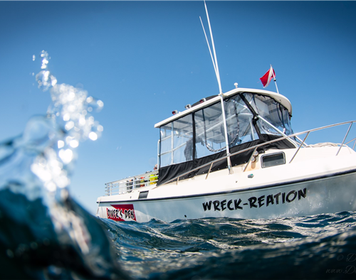 A small boat named Wreck-Reation in the water with a small wave coming at the camera