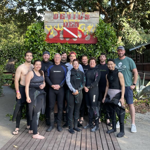 Group of adults in wet suits standing in front of a sign that says Devils Den with a scuba diving flag on it