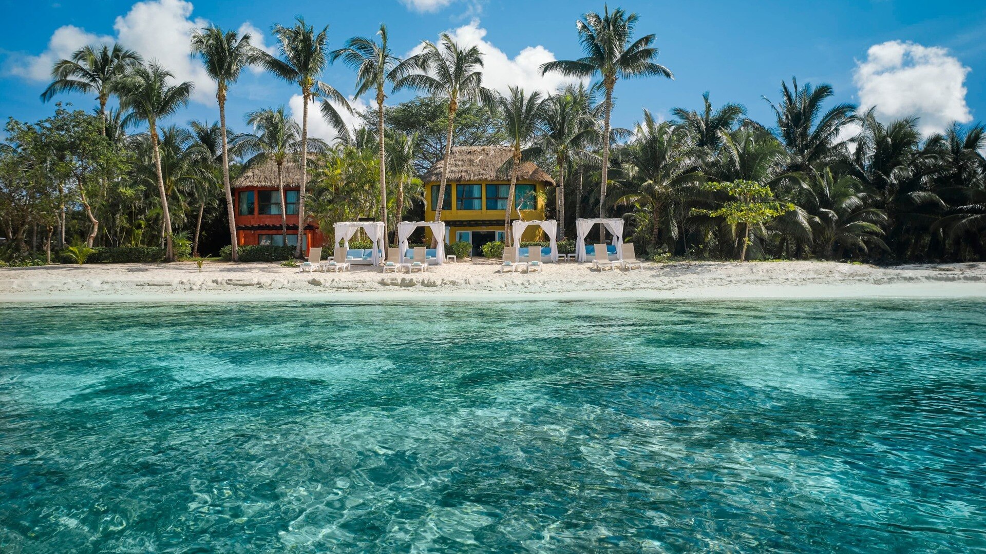 Beach with clear blue water. Lounge chairs and couches are set up under canopies